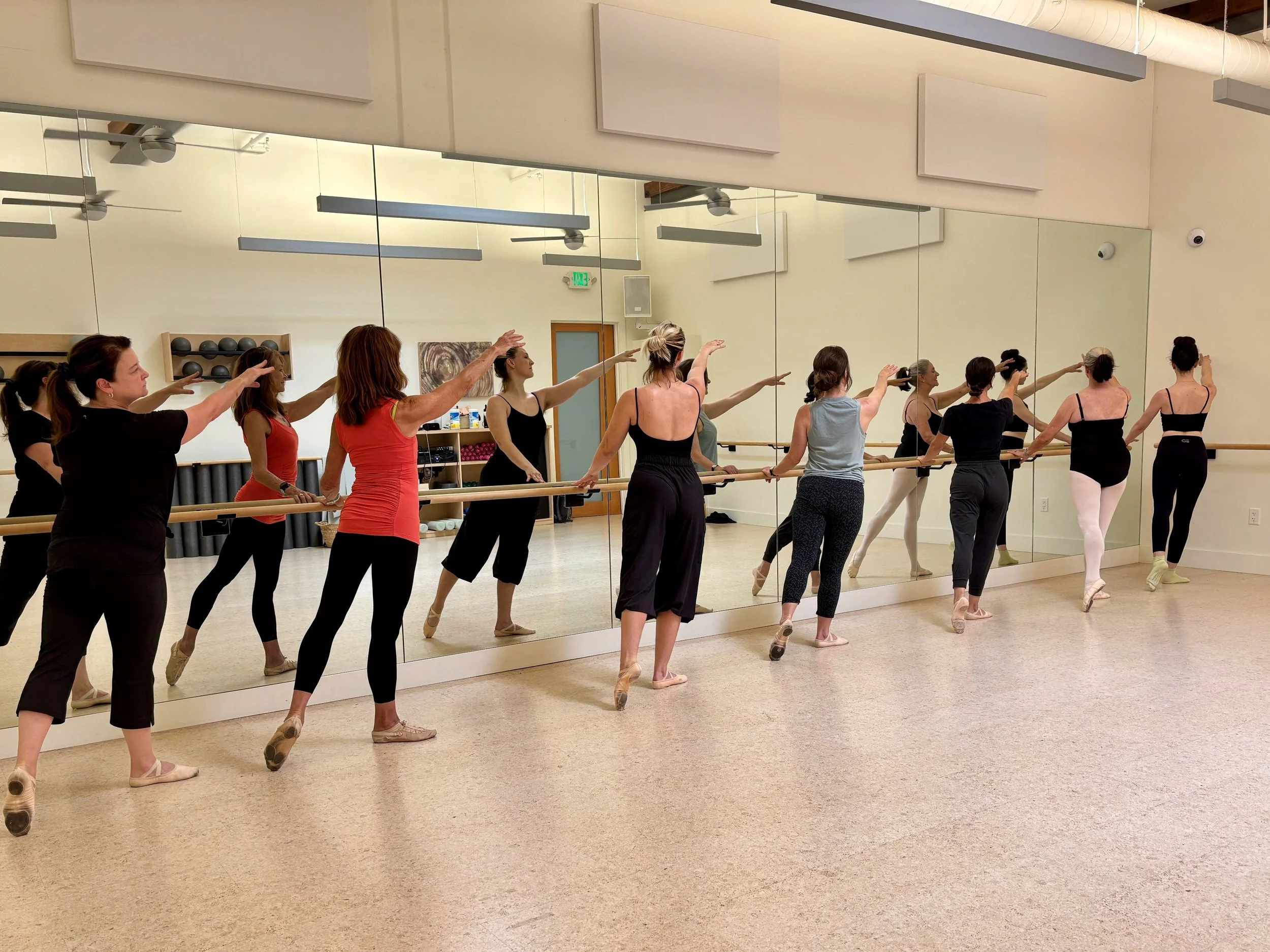 Adult dancers practicing together at a ballet barre in a mirrored studio.